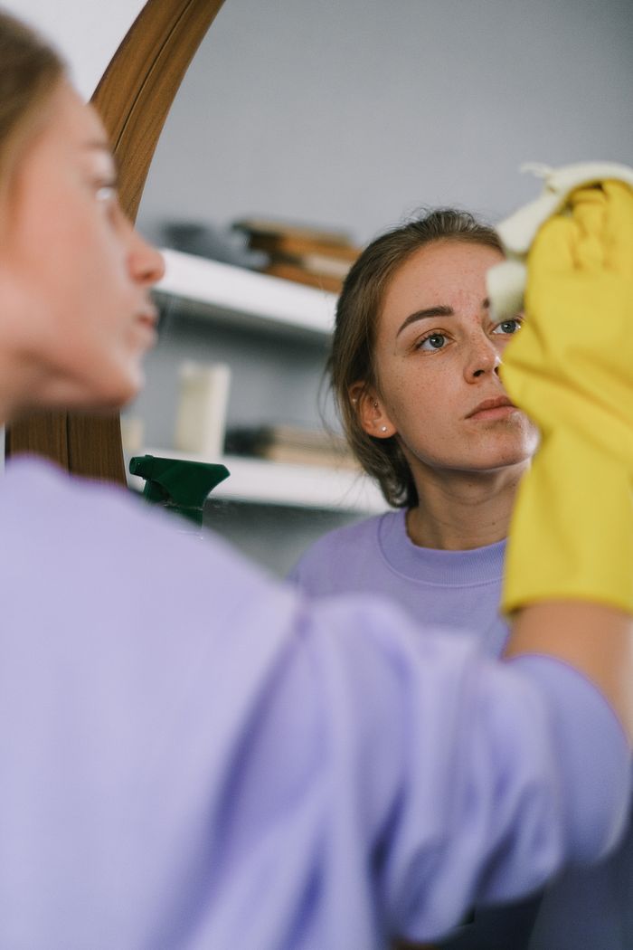 hero-img-02 Crop female in purple sweatshirt and latex gloves washing mirror in light apartment in daytime